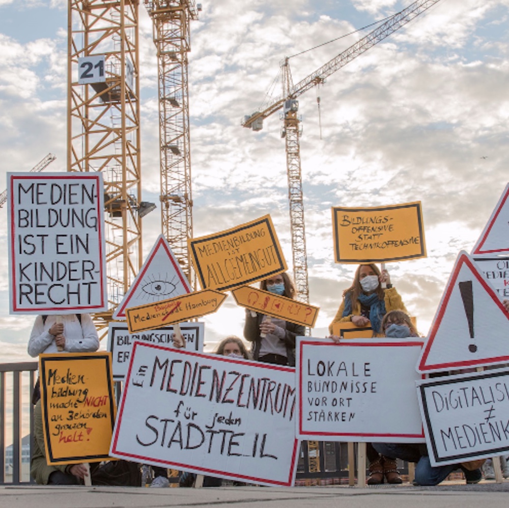 Foto von mit Forderungen und Slogans beschrifteten Schildern vor einem Baustellen-Hintergrund, z.B. "Medienbildung ist ein Kinderrecht", "Ein Medienzentrum für jeden Stadtteil", "Mehr Medienbildung für alle"