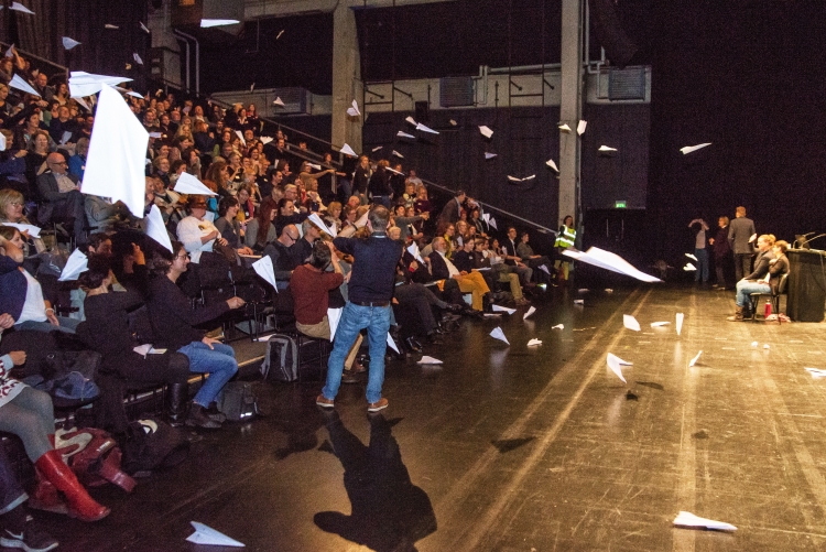 Foto: Menschen werfen Papierflieger von der Zuschauertribüne auf die Bühne