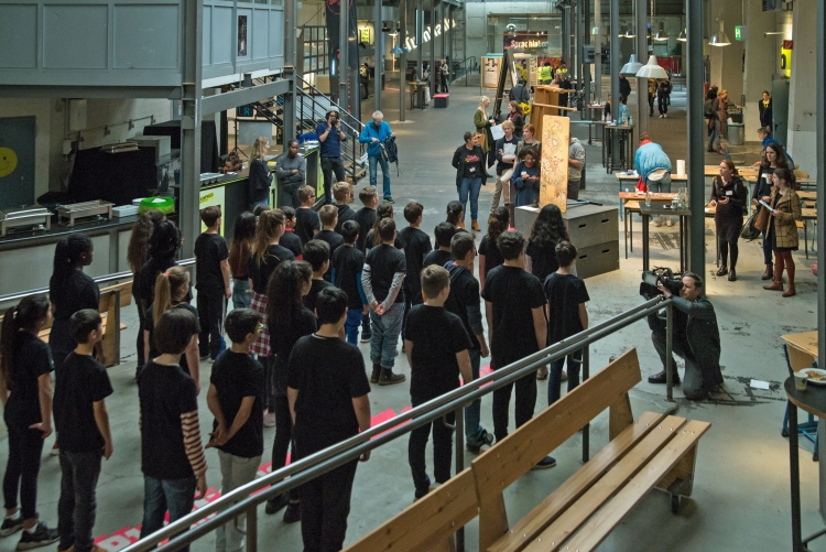 Foto: Schüler*innen in schwarzen T-Shirts in Reihe und nebeneinander stehend von hinten im Foyer