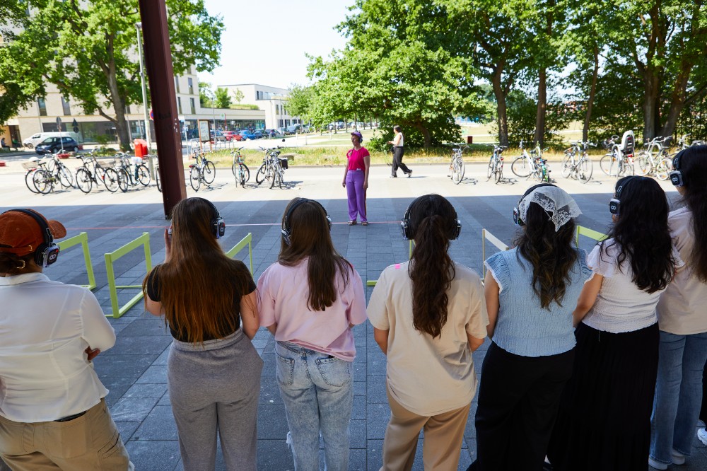 7 Jugendliche im Freien. Sie stehen mit dem Rücken zu uns vor einem Parkour und haben Kopfhörer auf.
