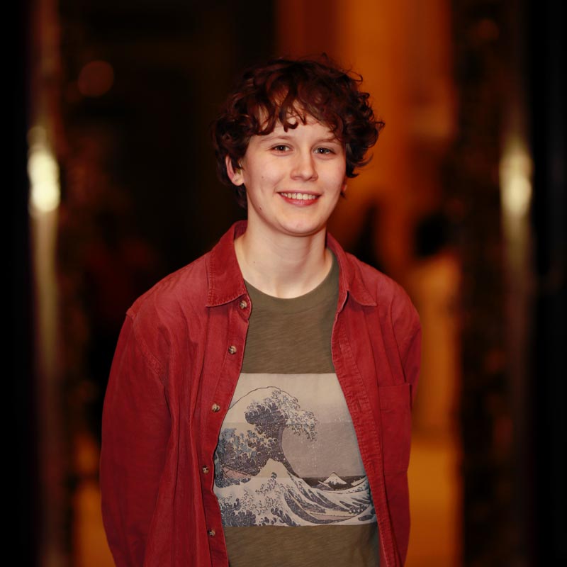 Porträt von Emilia:  lächelnde, weiße Person mit kurzen, lockigen Haaren